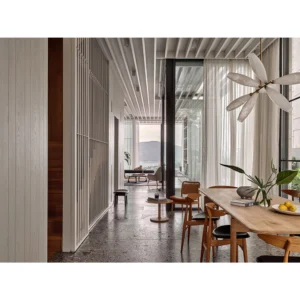 The image is a modern dining area with large glass windows offering a view of a mountainous landscape under a cloudy sky. Inside, there’s a wooden dining table with chairs, stools, and an armchair. The polished concrete floor contrasts with vertical wooden slats on one wall. Notably, a large chandelier hangs above the dining area.