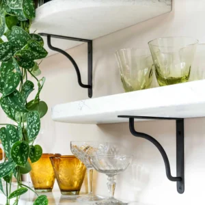 The image features Corston’s Bronze Kilburn Shelf Bracket supporting two white marble shelves. The top shelf holds a potted plant, while the bottom one displays amber-colored glasses and clear glass bowls. The black brackets create a modern aesthetic against the white wall.
