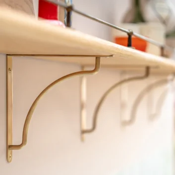 The image features an Antique Brass Kilburn Shelf Bracket by Corston. The bracket is polished and made of antique brass, showcasing an elegant curve and vintage design. It is mounted on a white wall, supporting a wooden shelf with various items on top. This high-quality bracket is ideal for interior design enhancements and functional shelving solutions. - Antique Brass Kilburn Shelf Bracket by Corston.